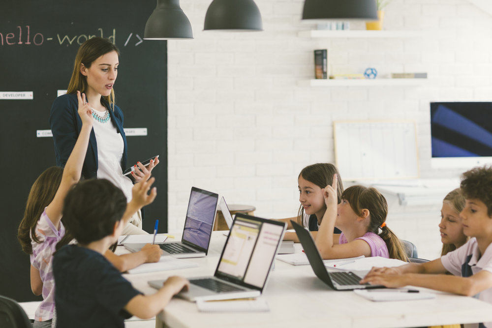 Group of children at coding class. They are sitting by the desk and using laptops. Their female teacher standing beside desk, holding digital tablet and asking question and children raising hands to answer.