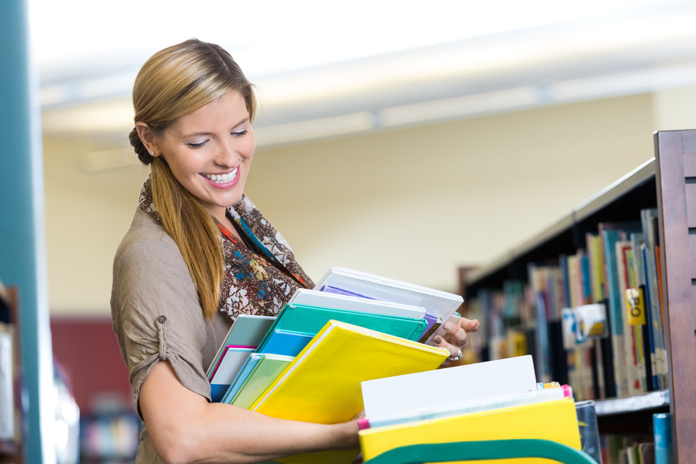 Mid adult Caucasian woman is librarian in elmentary school library. She is smiling while putting books away on bookshelves. She is pushing a rolling cart filled with children's books, and is wearing casual trendy clothing.