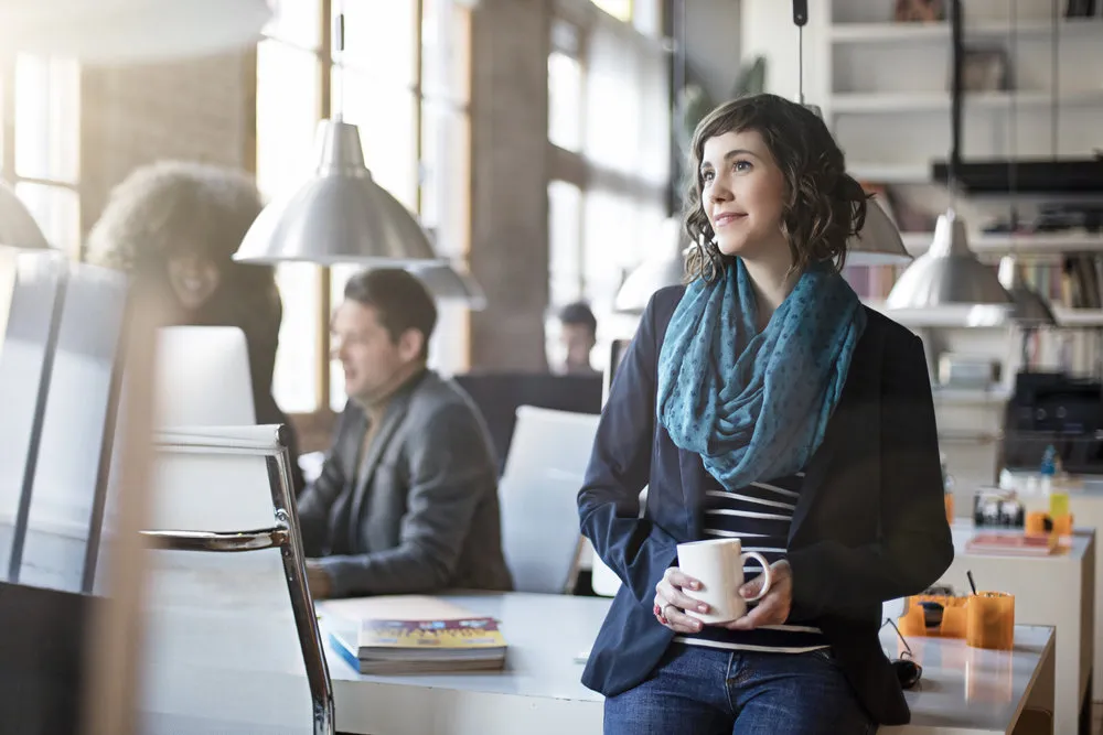 Portrait of smiling office worker relax having a coffee on a sunny day, her colleagues are working in the background.