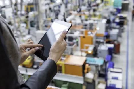 Horizontal color image of businesswoman - unrecognizable person - working with digital tablet in large futuristic factory. Woman standing on top of a balcony, holding touchpad and checking inventory of a manufacturing company on touchscreen tablet. Focus on businesswoman's hands holding black tablet, futuristic machines in background.