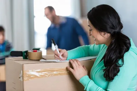 Mid adult Hispanic mother writes a to-do list as she and her family pack up to move to new home. Her husband and son are packing or moving boxes in the background.