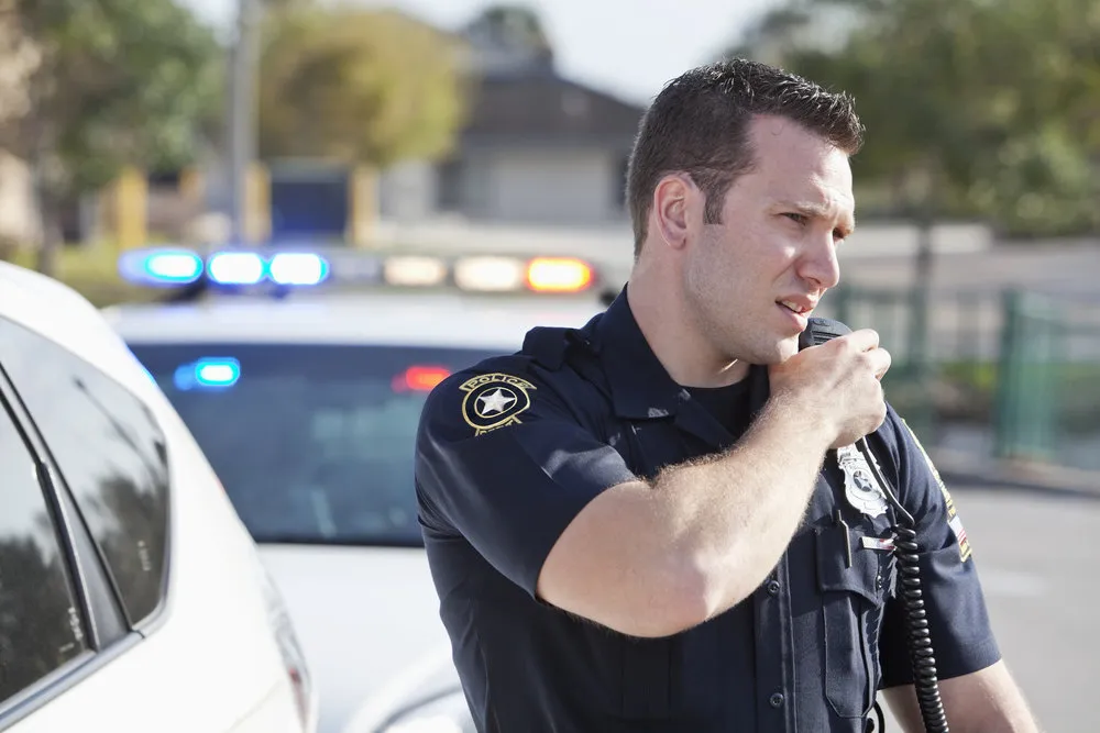 Male police officer (20s) standing outdoors, reaching for radio. Police car in background.