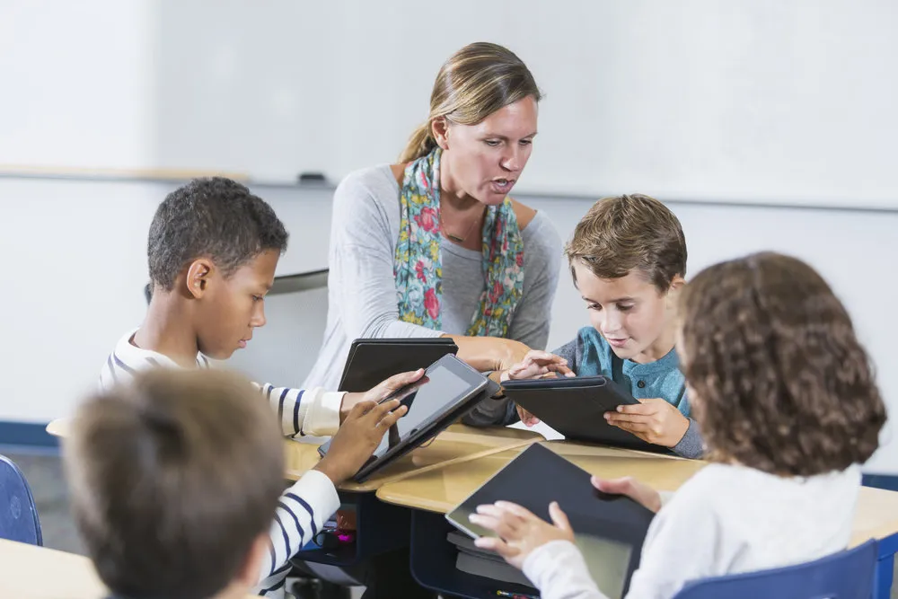 A teacher and a multi-ethnic group of elementary school children sitting around a table in the classroom, each one using a digital tablet. The woman is helping a boy use his.