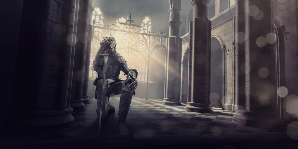 A medieval knight in full suit of armour kneels whilst holding his sword to his head in contemplation or prayer before a big battle. The knight is inside an ancient medieval stone building, which could be a castle or cathedral as the evening from the setting sun streams through the windows.