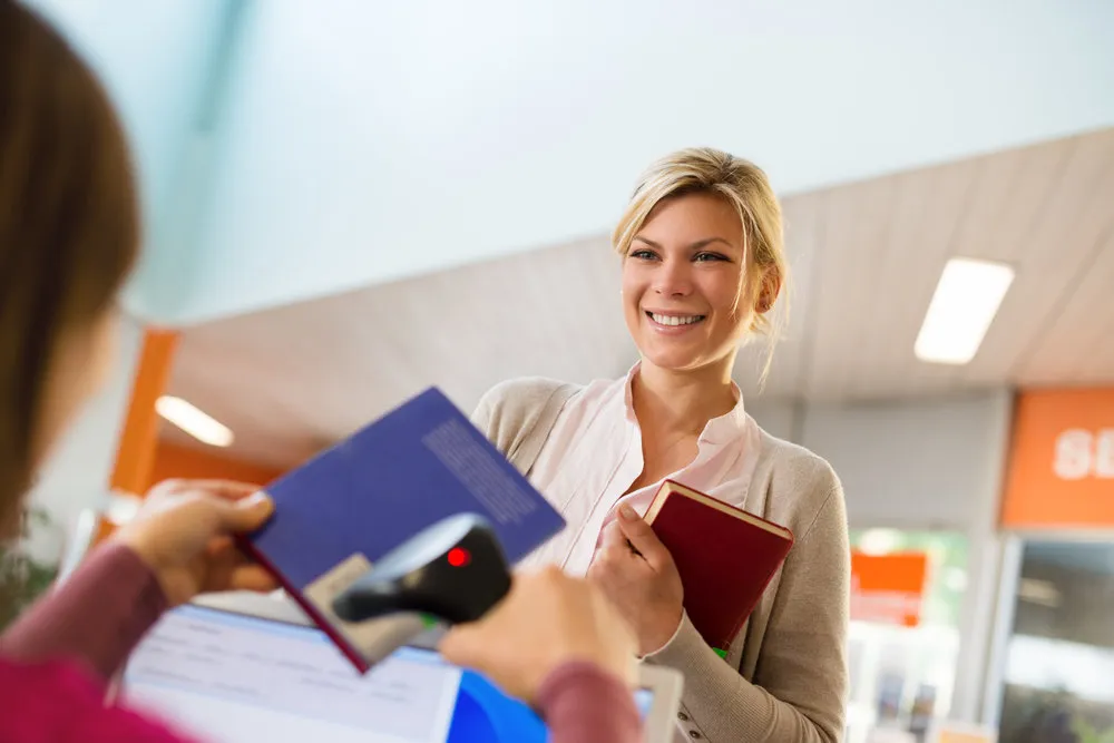 "Portrait of young female college student returning book to library, with librarian scanning barcode"