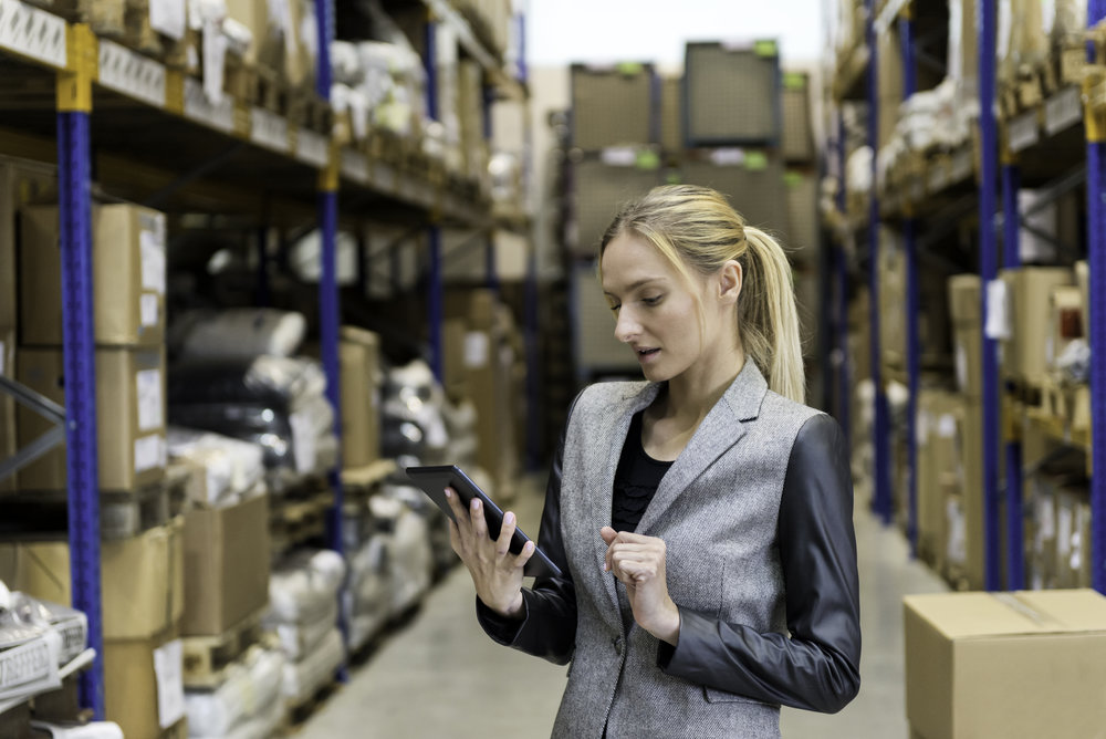 Portrait of a pensive young woman supervisor working on-line in warehouse. Young blond woman standing at distribution warehouse and wearing elegant suit. Industrial boss examining the stock. Large distribution storage in background with racks full of packages, boxes, pallets, crates ready to be delivered. Logistics, freight, shipping, receiving.
