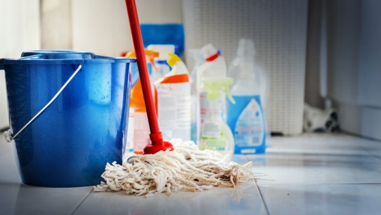 Closeup of unrdcognizable home cleaning products with blue bucket and a mop in front in sharp focus. All products placed on white and poorly lit bathroom floor.