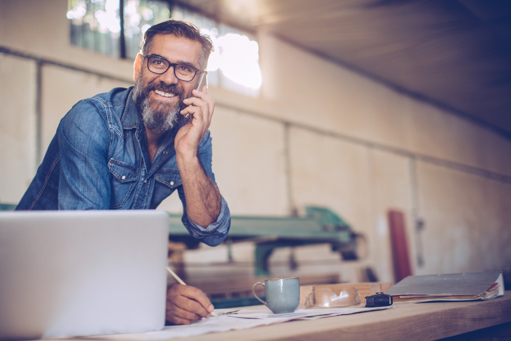 Carpenter in wood workshop using phone and working on project. Mature man in casual clothing. Space is full of working tools and wooden planks.