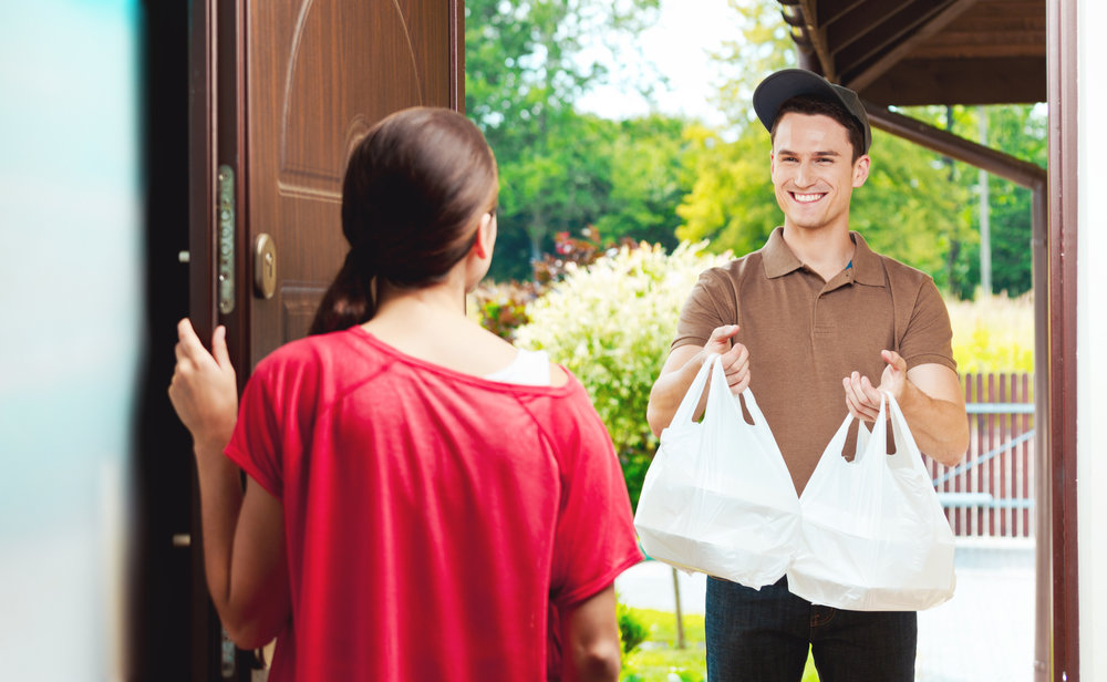 Young delivery man delivering chinese take away food for young woman, standing at the entrance door and holding two plactic bags in hands.