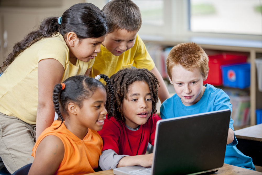 A multi-ethnic group of elementary age children are sitting in class playing an educational game on a laptop.