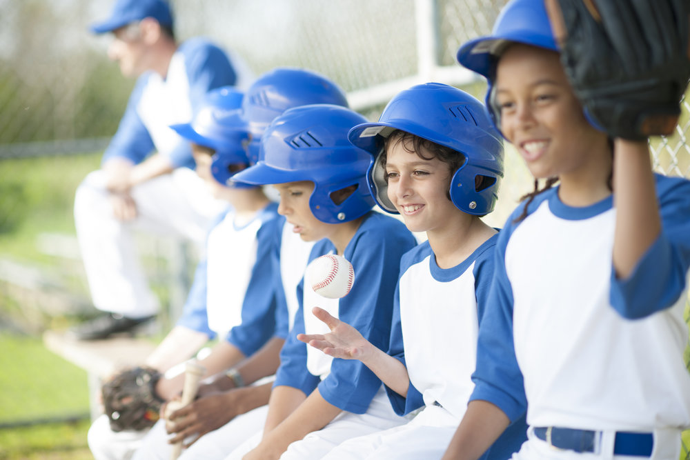 A multi-ethnic group of elementary age boys are sitting in the bench waiting for their turn to bat during the baseball game.